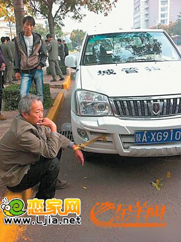 一輛寫著“城管執(zhí)法”字樣的白色汽車旁坐著一位白發(fā)蒼蒼的老漢，被一根黃色的繩子拴在車